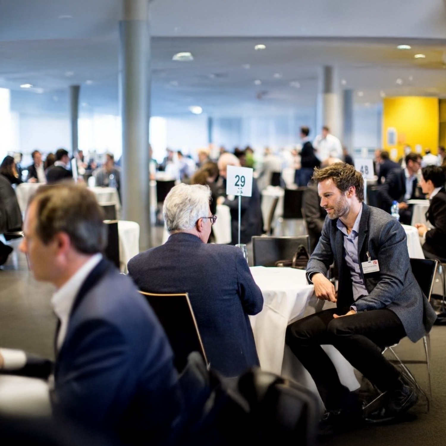 Professionals engaged in structured one-on-one business meetings at a large networking event or conference, with numbered tables in a spacious venue.